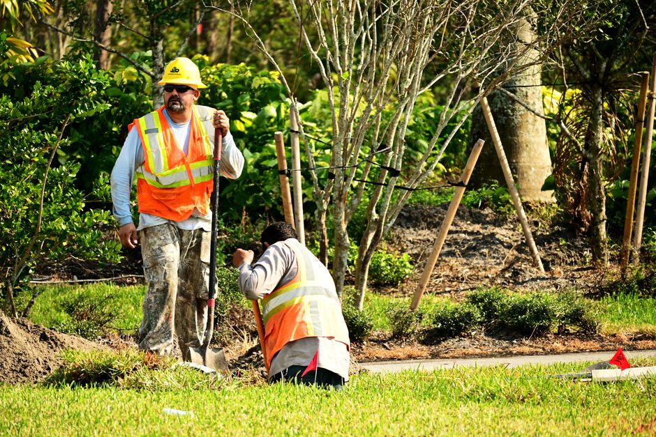 Two gardeners wearing high-visibility safety vests and protective helmets are engaged in garden maintenance work in a lush, well-kept outdoor space. One gardener, standing upright, holds a long-handle
