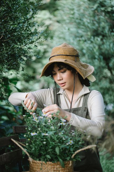 A young woman wearing a wide-brimmed straw hat and a beige long-sleeved top is engaged in garden maintenance, carefully tending to a potted plant or flowers amidst lush green foliage. She holds a smal