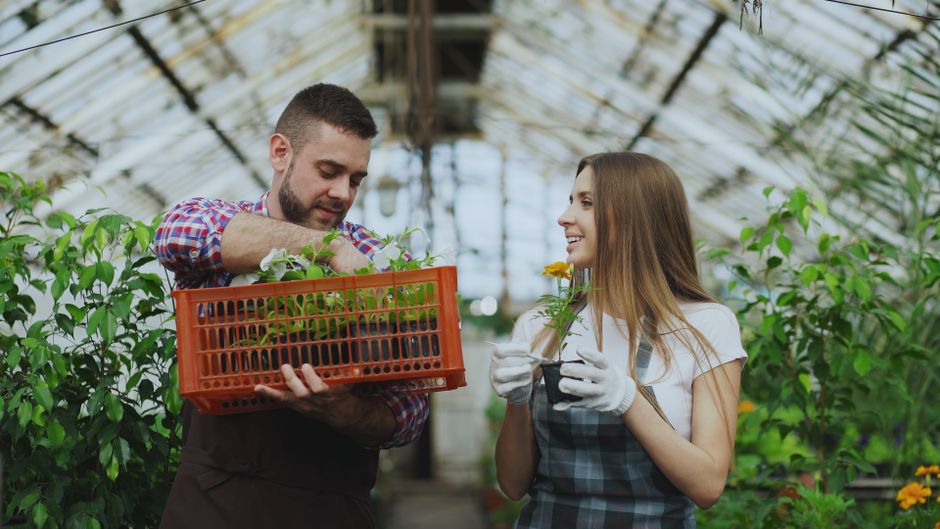A young woman wearing a wide-brimmed straw hat and a beige long-sleeved top is engaged in garden maintenance, carefully tending to a potted plant or flowers amidst lush green foliage. She holds a smal
